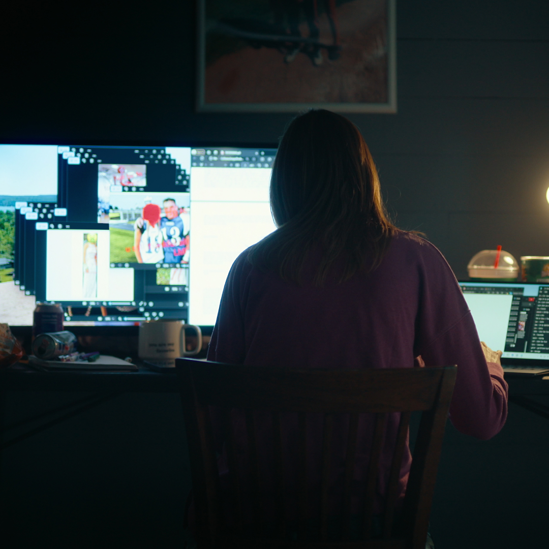 A woman facing away from the camera works on a desktop computer and laptop in a dark room with a desk lamp on, in 'Unknown Number: The High School Catfish' on Netflix.